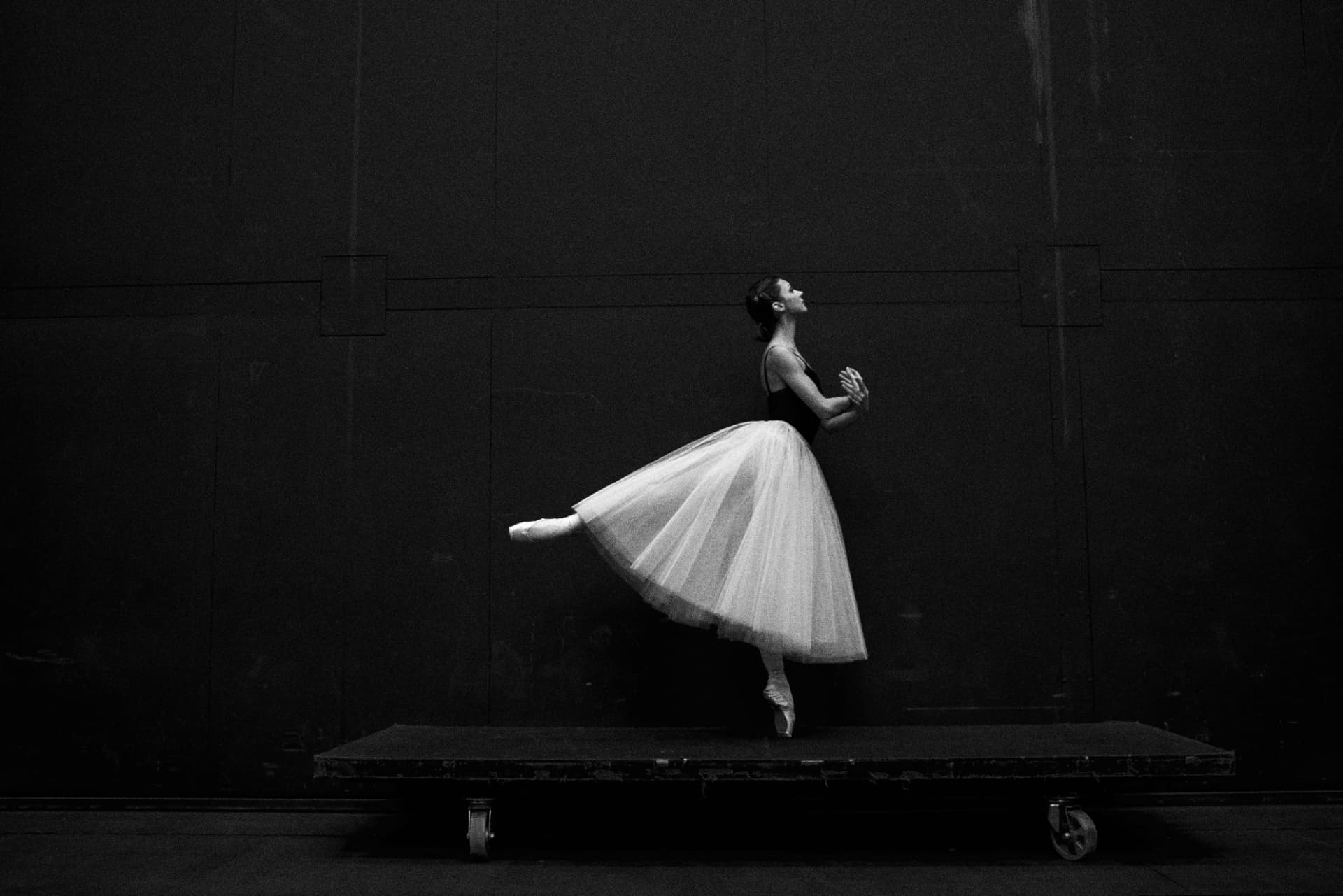 Bailarina en puntas y tutú blanco, de perfil, en equilibrio sobre una plataforma móvil con ruedas frente a un fondo oscuro, en blanco y negro.